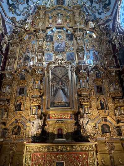 This image captures the highly ornate and gilded interior of a Baroque or Churrigueresque religious building in Tepotzotlán, Mexico. The scene is dominated by an overwhelming display of gold leaf, intricate carvings, and numerous religious artworks.
The walls and possibly part of the ceiling are densely covered with a lavish framework of gold, featuring elaborate scrolling motifs, foliage, and architectural elements. Embedded within this golden matrix are a multitude of small, framed paintings. These paintings depict various religious subjects, including portraits of saints, figures of Christ, and the Madonna and Child. Many of these artworks are set within rectangular, octagonal, or diamond-shaped frames.
Several small, sculpted figures, likely saints or angels, are also integrated into the golden decoration, often appearing within niches or as part of larger ornamental structures. The inclusion of multiple small mirrors within the gilded elements adds to the dazzling effect, reflecting light and enhancing the opulence.
A large, central painting is a focal point, featuring multiple figures, possibly depicting the Virgin Mary with Jesus and surrounding angels, all framed by an exceptionally detailed golden border. On the far right, a section of a dark red panel adorned with intricate gold patterns suggests further rich decoration. In the upper left corner, a portion of a ceiling or upper wall with a fresco in shades of blue and white provides a contrast to the prevailing gold.
The image presents a static view of this intensely decorated space, highlighting the architectural and artistic grandeur rather than any ongoing activity. The sheer volume of gold, the intricate craftsmanship, and the religious iconography evoke an atmosphere of profound reverence and splendor. No discernible text is visible in the image.