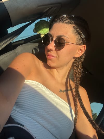 In the city of Stolnik, Bulgaria, a young woman with dark hair styled in braids adorned with silver rings sits in the passenger seat of a car. She wears round sunglasses and a white strapless top. A small, green plush turtle hangs from the car's interior. The setting sun casts a warm glow on her face and arm, creating a shadow across her chest, where a tattoo reading "She flies with her own wings" is visible. The car's seatbelt is fastened, and the window shows a glimpse of the outdoors with trees and a blue sky.