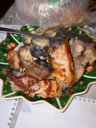 The image shows a plate of cooked fish. Some pieces of fish are lightly colored and appear to have been poached or steamed, while others are darker, suggesting they have been fried or baked. A metal serving utensil is partially visible, resting among the fish pieces. The plate is star-shaped with a green and yellow border, decorated with Christmas-themed designs, including stars, lanterns, and ornaments. A clear plastic bag and what appear to be various nuts are partially visible near the plate. The scene is on a white surface. There is no information in the image to confirm the location or city.