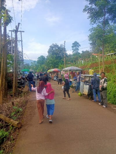 This image depicts a lively roadside market or gathering in a rural or semi-rural setting, likely in Kecamatan Pangalengan, Indonesia. The scene unfolds on a paved road surrounded by lush green trees and vegetation, with distant hills or mountains visible under a bright, partly cloudy sky, suggesting a clear morning or early afternoon.
Numerous people, dressed casually, are present, walking, standing, and browsing. Several informal market stalls, identified by colorful umbrellas (red, orange, green) and awnings, line the road. These stalls appear to be selling various goods, possibly food or local products, with some looking like mobile food carts, indicated by visible propane tanks. Motorcycles are also parked near some of the stalls.
In the foreground, two young women, both wearing hijabs, walk arm-in-arm away from the viewer. On the right side of the frame, a man in a patterned white and black shirt smiles directly towards the camera, standing near another man in a dark hoodie who is looking towards the market stalls. Utility poles and power lines run along the left side of the road, and some cut logs lie on the ground. The overall atmosphere appears relaxed and communal, set against a backdrop of natural beauty characteristic of highland regions. No visible text is clearly discernible.