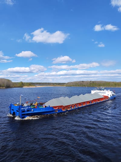 A barge filled with gray gravel is moving across a wide, dark blue river under a bright blue sky with scattered white clouds. Trees line the opposite bank. The barge is blue and orange with white accents. "ОКСКИЙ-45 И" is printed on the side of the barge. The photograph appears to be taken in a rural or natural environment, not within the city of Edoma, Russia.