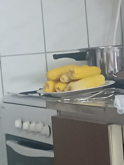 The image displays a close-up, slightly blurry view of a kitchen scene. In the foreground, a white stove with black knobs is visible. Above the stove, a white plate holds several cooked ears of corn. Behind the corn, a stainless steel pot with a black handle is partially visible. The background consists of white tiled walls. The overall impression is that of a domestic kitchen setting. The image does not contain any identifiable landmarks or specific architectural features that would allow for a precise determination of its location within Ashgabat, Turkmenistan.
