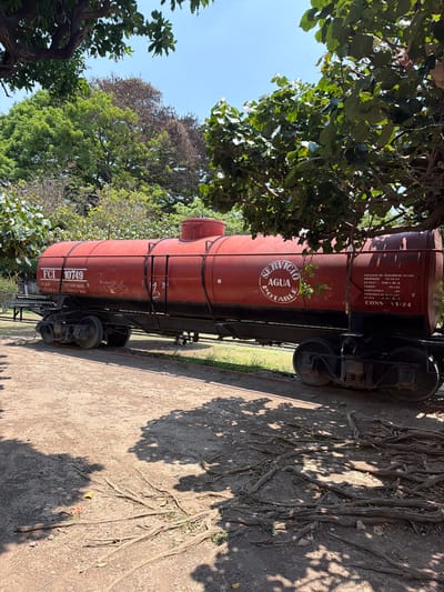 The image depicts a red vintage water tank car labeled "FCI 10749," which is standing on a section of track in a park-like setting. The tank car is marked with "Servicio Agua Potable" indicating it once served as a potable water supply. Trees surround the tank car on both sides, and a network of roots covers the ground alongside the tracks. Shadows from the trees create patterns on the ground. The OCR reveals more details such as information about the car's safety valves, testing dates (1964), and weight capacities. Based on other publicly available data, this scene is likely located in the city of Cuautla, Mexico, where similar vintage railroad equipment has been installed as historical displays in parks and recreational areas.