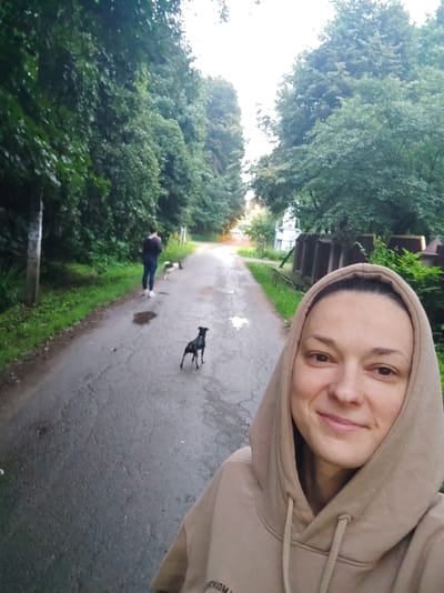 A woman in a tan hooded sweatshirt is taking a selfie outdoors on a wet, paved road lined with trees. She is smiling and her face is in the foreground. In the background, a person in a dark jacket and blue jeans is walking a dog. A second, smaller dog is walking on the road further ahead. The environment appears to be a residential area with houses and fences visible in the distance. The scene is captured during daylight, likely in the morning or evening given the lighting. The location is identified as Alabino, Russia.