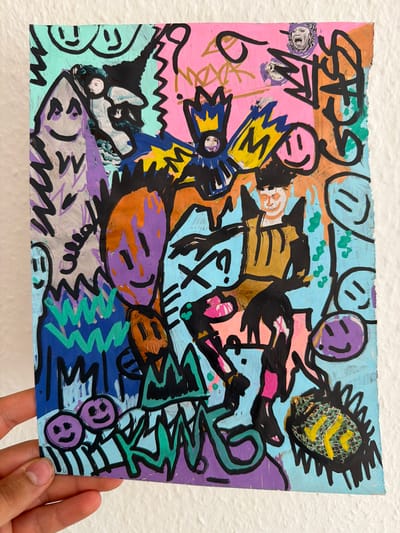 This is a photograph of a mixed-media artwork featuring a vibrant and chaotic composition. The subjects are abstract and cartoonish figures, some resembling faces with simple lines for eyes and mouths. There are also elements that could be interpreted as abstract objects, like a spiky, textured form in the upper right corner with yellow markings, and what appears to be a crown-like structure in yellow and blue on the left.

The artwork is dense with black marker lines over colorful paint, including pink, blue, orange, purple, gold, and green. The style is reminiscent of graffiti or street art, with bold outlines and energetic scribbles. Some areas show a distressed texture, suggesting the use of torn paper or textured paint. There are small photographic cutouts of what appear to be classical busts embedded in the artwork.

The overall mood is energetic and perhaps playful or aggressive due to the strong lines and clashing colors. No specific activity or event is clearly depicted; it seems to be a collection of abstract forms and figures. There are no discernible environmental cues for time of day or weather, and the setting is neutral, as the artwork is presented against a plain white background. There is no visible text in the artwork itself. The photograph is taken in Nienburg/Weser, Germany, with a hand holding the bottom right corner of the artwork.