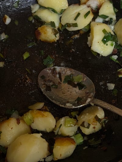 The image shows a close-up, overhead view of a dark, possibly cast-iron skillet filled with cooked potatoes and chopped green onions. Some of the potatoes appear golden brown and slightly crispy on the edges, while others are pale yellow. The green onions are scattered throughout, with some pieces wilted and others still bright green. A silver spoon with food residue on it rests in the skillet, angled from the top right towards the bottom left. The skillet appears to be on a dark surface, but the surroundings are not clearly visible. There are no people or animals present in the image. The scene depicts a finished dish or food preparation in progress. This scene is not located in Satino-Russkoe, Russia, as the image does not contain any elements that would indicate a specific geographical location.