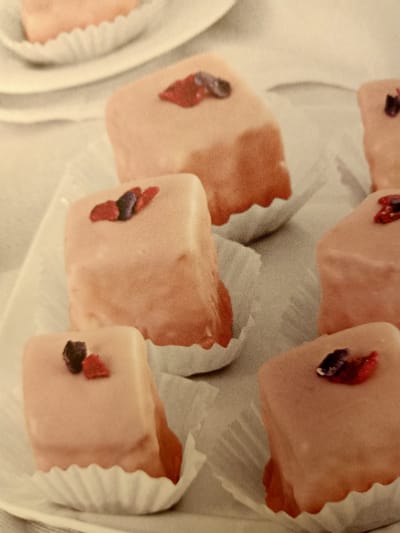 The image displays a close-up view of several small, square-shaped cakes, possibly petit fours, arranged on a white surface. Each cake is topped with a pinkish glaze and adorned with small pieces of what appear to be dried berries, one red and one dark purple. The cakes are presented in white paper liners. The background is softly blurred, suggesting a shallow depth of field. There are no people or discernible elements of the city of Rumaithiya, Kuwait, in the image.