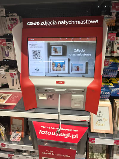 The image shows a red and white photo kiosk labeled "CEWE" in what appears to be a retail store. The kiosk screen displays "CEWE zdjęcia natychmiastowe" (CEWE instant photos) and a QR code for transferring photos from a smartphone. Various products, including USB cables, batteries, and picture frames, are visible on shelves in the background. The kiosk has slots for USB and SD cards, as well as a printer output. The overall environment suggests a consumer electronics or department store. The kiosk is positioned in an aisle with other retail displays. There is no indication of the specific location within the city of Brzeg, Poland.