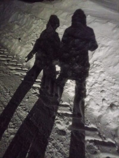 The image depicts a nighttime outdoor scene in the city of Старый Шагирт, Russia. Two human figures are visible as dark silhouettes against a snow-covered environment, illuminated from behind by an unseen light source that casts long, distinct shadows towards the viewer. The figure on the left is smaller, with its right arm slightly extended forward. The figure on the right is taller and stands upright. Both figures appear to be wearing hooded garments. The ground is uneven, entirely covered in snow of varying depths and textures, with visible footprints or depressions in certain areas. No other distinct objects or specific actions are discernible.