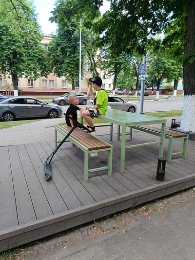 Two young boys are seated on a picnic table and bench in Gubkin, Russia. The boy on the left is wearing a black t-shirt and shorts, while the boy on the right is wearing a bright green t-shirt and patterned shorts. They appear to be interacting, with the boy on the left looking up at the boy on the right. A scooter lies on the wooden platform in front of the bench. The picnic table and benches are made of wood and painted a light green. In the background, there are trees, a street with cars, and a residential building. The area appears to be a public space, possibly a park or recreational area.