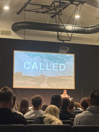 The scene is indoors, seemingly in an auditorium or large meeting hall. A projection screen displays the word "CALLED" in large, light-colored letters, with the subtitle "CALLED TO WALK TOGETHER". The screen also shows a blurred image of a beach or coastline. A projector is attached to the ceiling via a mount. In the foreground, a crowd of people is seated, facing the screen. A speaker is faintly visible at the front of the room, near the projection screen. The lighting is dim, with a spotlight illuminating the speaker and screen. The location appears to be a generic auditorium and cannot be specifically identified as being in Springfield, United States of America. Other visible objects include a metal support structure and a large ventilation pipe running along the ceiling.