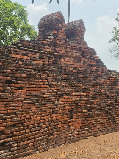 An upward-angled view captures a large, ancient brick wall structure, likely part of a historical ruin in Phra Nakhon Si Ayutthaya City Municipality, Thailand. The wall dominates the frame, showcasing layers of weathered, reddish-brown bricks. Many bricks exhibit discoloration, dark stains, and signs of erosion, indicating significant age.
On the left side of the image, the brick structure tapers upwards, featuring distinct tiered or corbelled sections that suggest an architectural detail of a tower or building edge. Green foliage from trees is visible on the far left and at the bottom left, partially obscuring the base of the structure and framing the top against a bright, clear sky with soft white clouds. The right side of the main wall extends into the distance, meeting a patch of sandy, earthy ground covered with scattered dry leaves.
No human activity is depicted. The scene suggests a bright, clear daytime. There is no visible text. The overall impression is one of ancient history and architectural resilience against time.