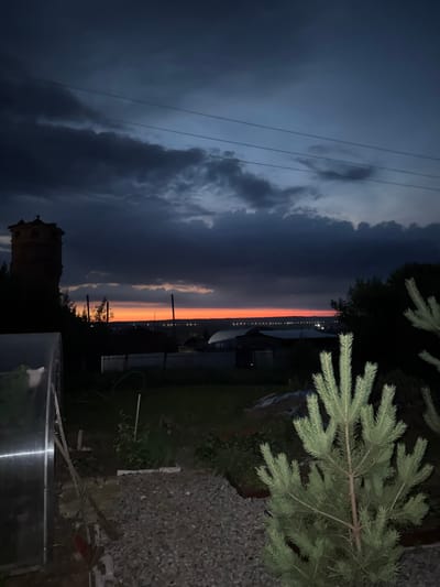 The image depicts a nighttime scene in Malmyzh, Russia, during what appears to be twilight or dusk. The sky is dramatic, with dark, stormy clouds dominating the left side of the frame, gradually giving way to a vibrant band of orange and red on the horizon, indicating the setting or rising sun. In the distance, a line of lights suggests a settlement or town.
In the foreground, to the right, is a branch of a coniferous tree, likely a fir or spruce, with its needles sharply defined. Below the tree, the ground is covered with gravel. To the left, silhouetted against the sky, is a dark, brick structure, possibly an old tower or building. In the lower right corner, a portion of a transparent structure, possibly a greenhouse or similar enclosure, is visible.
The overall mood is somber due to the dark clouds, but the colorful horizon adds a touch of dramatic beauty. The presence of power lines across the sky further grounds the scene in a rural or semi-rural setting. There is no visible text in the image. The activity taking place is simply the natural progression of the day into night.