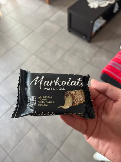 A hand is holding a package of Markolatu Wafel Roll with tahini cream. The package is black with white writing. The writing on the package says "Markolatu Wafel Roll, Me Krema Taxini with Tahini Cream." The hand is holding the package in front of a white tiled floor. The coordinates (40.627, 24.575) correspond to a location in Limenaria, Greece. This location is likely an indoor space, potentially a residential home. The environment is typical of a residential setting, with a tiled floor and a hand holding a food product. The image was likely captured by a person to document the product.