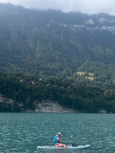 This scene is located on Lake Brienz, adjacent to the mountainous landscape of Bönigen, Switzerland. A single individual is seated on a light-colored stand-up paddleboard (SUP) in the foreground. The person wears a white baseball cap, a blue life vest, and pink shorts, holding a dark cylindrical object with both hands in front of their face. A paddle is visible resting on the SUP, along with a dark bag secured to the board's front. The lake water exhibits a distinct turquoise hue. In the background, steep, densely forested mountains rise directly from the lake, featuring a prominent light-colored rock face. Chalet-style houses with dark roofs are sparsely distributed on the lower forested slopes, interspersed with small green clearings. The sky is overcast.