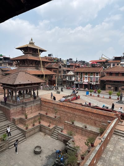 The image depicts a view of Patan Durbar Square in Lalitpur, Nepal. It's an elevated perspective of the square, showing brick buildings with multi-tiered roofs, intricate woodwork, and traditional Newari architecture. People are walking around the square, some sitting on the raised platforms of temples or structures. A few individuals are visible near the bottom right, walking on steps. In the upper part of the frame, shops with signs are visible, indicating a tourist presence. The sky is partly cloudy. The scene captures daily life and the architectural heritage of Patan Durbar Square.