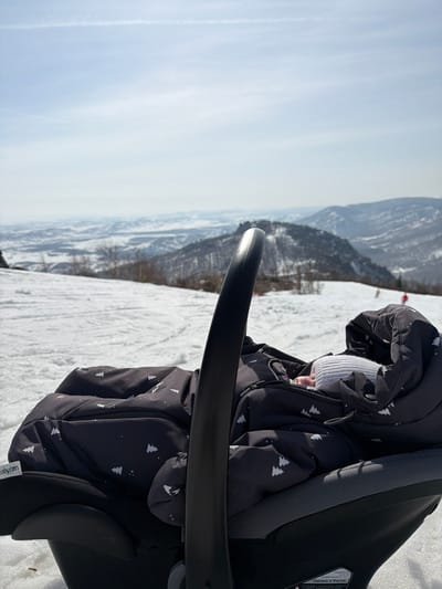 A light-colored infant, wearing a white knitted hat, sleeps enclosed in a black and white patterned winter bunting bag within a black and grey infant car seat. The car seat, featuring a "babyten" tag, rests directly on snow-covered ground on a mountain slope. The perspective is low, looking past the infant and carrier's black handle towards a vast, snow-laden mountain landscape under a bright, clear blue sky with a faint contrail visible overhead. Distant snow-capped peaks and hills, some sparsely forested with bare trees, extend to the horizon. The foreground and mid-ground consist of a groomed, snow-covered slope, possibly a ski run, with subtle track impressions. Small, distant figures, likely skiers, are discernible further down the slope. The scene is located in Zelenaya Polyana, Russia.