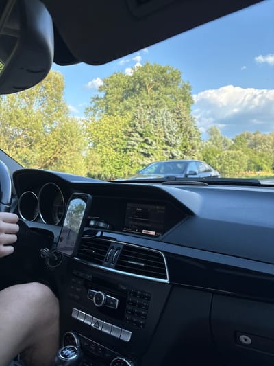 The image shows the interior of a car, looking out through the windshield. The car appears to be parked or driving slowly on a road bordered by trees and vegetation. A dark-colored car is visible in front of the vehicle. The dashboard of the car has a central display screen showing navigation and audio information, and a smartphone mounted on a holder. A person's arm and hand are visible resting on the steering wheel. The environment suggests a suburban or rural setting under a blue sky with scattered clouds. The location is identified as Iffezheim, Germany.