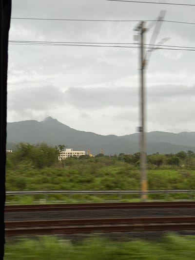 The image depicts a view from a moving vehicle, likely a train, looking out at a landscape. In the foreground, a blurry section of train tracks runs horizontally across the frame. Beyond the tracks, a dense expanse of green foliage, possibly trees and bushes, stretches across the midground. A pale, almost white building with multiple windows is visible in the distance, situated amongst the greenery. Behind the building, a large, dark mountain range rises, its peaks shrouded in mist or clouds. The sky is overcast with a pale gray cloud cover. A blurry, vertical pole, possibly a utility pole, is present in the midground, partially obscuring the view of the landscape and sky. The overall impression is one of movement and a somewhat muted, possibly humid, atmosphere. This scene is consistent with the views often seen from trains traveling through the Palghar district of Maharashtra, India, which is characterized by its rolling hills, lush vegetation, and scattered settlements.