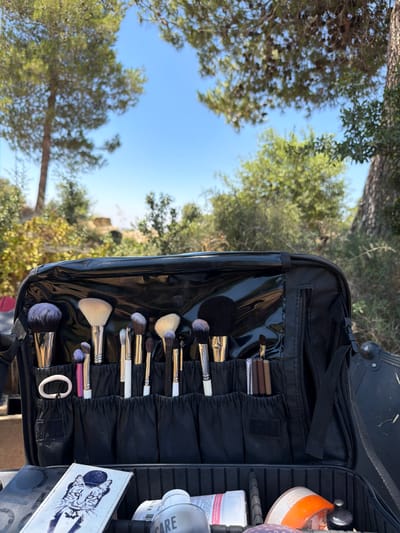 In Abu Gosh, Israel, a black makeup case is open, showcasing a variety of makeup brushes neatly organized in individual slots. The brushes have different sizes and colors, with some featuring black and silver handles and others white. Some brushes have purple tips. In the bottom compartment of the case, various makeup products are visible, including a white bottle with a pink label, a clear bottle, and a product with an orange lid. A white card with a drawing of a cat wearing a hat and bowtie is also placed in the bottom of the case. The case is positioned outdoors, with a background of green trees and a clear blue sky, suggesting a natural, possibly arid environment. The sun is shining brightly, casting shadows.