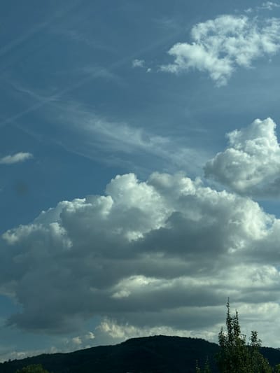 The image shows a landscape scene with a dark, forested hill in the foreground under a blue sky filled with white and gray clouds. The clouds are varied in texture and density, with some appearing fluffy and bright, while others are darker and more ominous. There are thin, wispy clouds and contrails visible higher up in the sky. A tree with dark green foliage stands on the right side of the frame, partially obscuring the hill. The overall impression is of a somewhat overcast or changeable weather day. The location is Mirovo, Bulgaria.