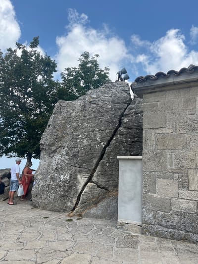 In the city of San Marino, San Marino, on a stone-paved ground, a large boulder with a deep fissure down its center is situated next to a stone building with a tiled roof. Two people, a man in a white t-shirt and denim shorts carrying a white plastic bag, and a woman in a coral-colored dress, stand near the boulder. Further behind them, another person is partially visible. Above the boulder, two spotlights are mounted on a rustic metal structure. The sky above is a vibrant blue with scattered white clouds. The scene is set outdoors, with trees providing a natural backdrop.