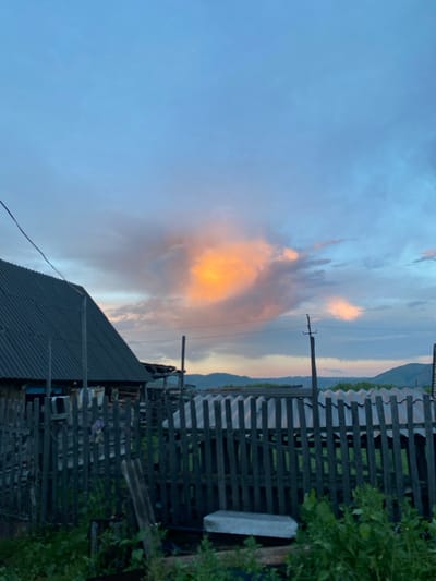 The image depicts a rustic rural scene with a prominent overcast sky featuring vibrant patches of orange and pink clouds, indicative of sunset or sunrise. In the foreground, a weathered wooden fence runs horizontally across the frame, partially obscuring the view of a modest dwelling with a dark, corrugated roof on the left. The house appears to be constructed from wood, and there are some indistinct structures and debris visible behind the fence. The background reveals rolling hills and a sparse landscape, suggesting a location outside of a densely populated urban area. A utility pole with wires is visible near the center of the image. The overall impression is one of a tranquil, natural environment within a rural settlement, possibly in Maloarslangulovo, Russia, given the description of the location. There are no people or animals visible in the frame. The vegetation in the foreground is dense and green.