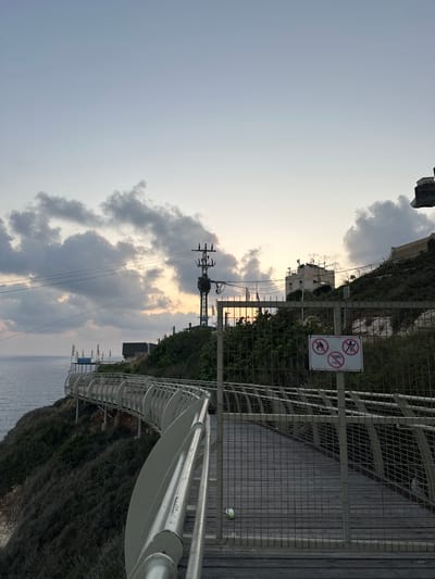 The image captures a scenic coastal walkway in the Mate Asher Regional Council, Israel, at dusk. The walkway, constructed of wood and metal railings, curves along a cliff overlooking the Mediterranean Sea. The sea is calm and a soft gradient of light appears in the sky, suggesting the sun has recently set or is about to rise. Dark clouds are scattered across the sky, with hints of orange and yellow light peeking through them.
On the cliffside, dense green vegetation covers the slope, interspersed with rocks and some structures. A tall utility pole with multiple wires stands prominently on the hillside, indicating proximity to infrastructure. Further up the hill, buildings and antennas are visible. A wire fence runs along the path, and a signpost displays three circular prohibition symbols: one with a flame, one with a person walking, and one with a bicycle. The walkway appears empty, with no people present. A discarded plastic bottle is visible on the wooden planks of the walkway, partially obscured by the fence. The overall atmosphere is serene and tranquil, with a touch of urban development meeting nature.