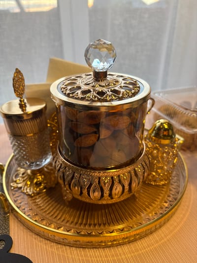 A close-up shot shows an ornate, gold-colored serving tray with several decorative items. At the center is a glass jar filled with almonds, topped with a clear, faceted crystal knob. The jar sits on a detailed gold base. To the left, a tall, slender gold and glass container stands, also topped with a decorative gold element. To the right of the central jar is a small, intricately detailed gold dispenser, likely for spices or sugar. The items are arranged on a textured gold tray. In the background, a sheer white curtain is visible, with sunlight filtering through, suggesting an indoor setting. There are no people or animals present. The scene is set on a textured peach-colored surface. The exact location in Dolche Vita KP, Russia, cannot be determined from the visual information provided.