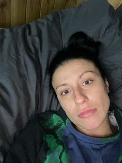 The media file contains a close-up, overhead shot of a person lying on a dark, textured surface, possibly a bed or couch. The person, identified as female with dark hair pulled back, is looking upwards towards the camera. Her face is the primary focus, with her eyes open and brown. She is wearing a dark hooded sweatshirt with a green and blue abstract pattern. The lighting suggests an indoor setting, possibly with soft, ambient light. There are no visible objects other than the person and the surface they are lying on. The background shows wooden planks, likely part of the wall or furniture. The scene appears to be a candid moment, with the person appearing relaxed or contemplative. There is no visible text in the image. The location context provided is Сарженка, Russia, but there are no specific visual cues within the image that confirm this location. The time of day and weather are not discernible from the image.