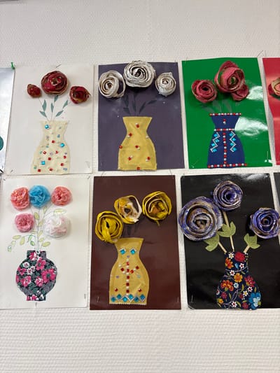 Six handmade art projects are displayed in a grid on a white, textured wall in Novosergievka, Russia. Each project depicts a vase holding flowers. The vases are constructed from various materials, including paper cutouts, felt, or patterned fabric, and are embellished with beads, sequins, or painted designs. The flowers are crafted from rolled paper, tissue paper, fabric, or dried organic materials, exhibiting a range of colors and textures. Each individual artwork is rectangular and secured to the wall with visible pushpins, primarily at the corners. Partial views of additional art pieces are visible on the left and right edges of the frame. The lighting is uniform, illuminating the artwork and the wall texture.