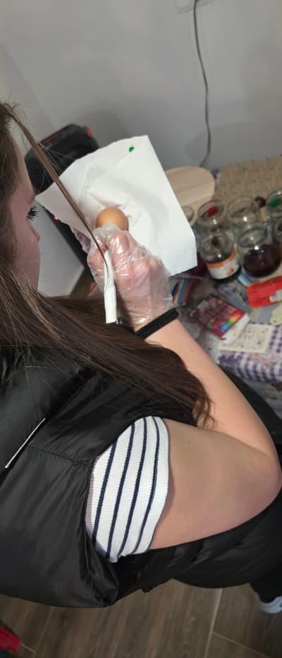 A person with long brown hair is dyeing a strand of their hair. They are wearing a black garment and striped socks. Their hand is covered in a clear plastic glove and they are using a white applicator. The hair strand lies on white paper with a brown egg on it. In the background are glass jars and containers on a wooden surface next to a wall. The location is in Novachene, Bulgaria.