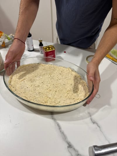 This image depicts a kitchen scene in Cidade da Maia, Portugal. A light-skinned individual, wearing a dark blue t-shirt and light-colored lower garments, is positioned over a white marble countertop with grey veining. The individual's forearms and hands are visible, actively holding a large, oval, transparent glass baking dish. The dish contains a uniform layer of light beige, granular substance, interspersed with small dark green flecks, consistent with seasoned breadcrumbs or a similar topping.
On the countertop, behind the baking dish, are several objects: a white bottle with a red label, a smaller dark bottle, and a red cylindrical tin with a gold lid, labeled "PIMENTÓN PICANTE" (Spicy Paprika) and featuring a chili icon. A small red substance residue is present on the countertop near the tin. To the right, a gold-colored metal ring and a folded, multi-colored (green, yellow, white) kitchen towel are visible. A silver kitchen utensil is partially visible at the bottom right. The individual's left wrist displays a red string bracelet, and a small, indecipherable tattoo is present on their right forearm.