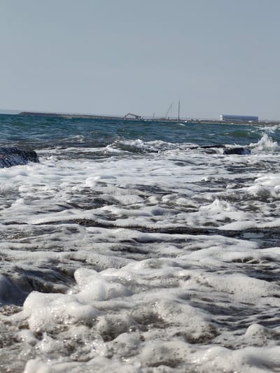 The image depicts a rocky shoreline with waves crashing onto the shore, creating white foam. In the background, there is a long breakwater with construction equipment, including cranes, visible. Further in the distance, a large building with a blue roof can be seen. The sky is clear and blue, suggesting a sunny day. The location is identified as a section of the shoreline in the city of Saiyn, Kazakhstan, likely a coastal area or a port under development. There are no people visible in the image. The scene is dominated by the natural elements of the sea and rocks, juxtaposed with the industrial presence of the construction site.