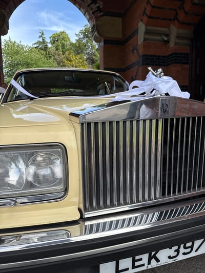 The image shows a close-up of the front of a light yellow Rolls-Royce car, adorned with white ribbon and a silver hood ornament. The car's grille and headlights are prominently visible. The vehicle is parked outdoors, under an archway made of red brick with black accents. Through the archway, green trees and a blue sky with white clouds are visible, suggesting a sunny day. The license plate of the car reads "LEK 397" and includes the inscription "Jack Barclay Limited". The scene appears to be set in front of a building, possibly a historic one, given the architecture. The overall impression is that of a formal occasion, likely a wedding or a special event. The location is the city of Hart, United Kingdom.