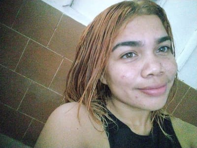 A woman with light brown hair, wet and styled straight, is photographed from the chest up. She is wearing a black tank top. Her skin is light-toned with a few blemishes visible on her face. She has brown eyes and full lips. The background consists of a tiled wall, likely in a bathroom, with a white surface at the top right. The overall lighting is dim and grainy, suggesting the photo was taken indoors with artificial light. No specific landmarks or indications of the city of Punta de Piedras, Venezuela are present in the image.