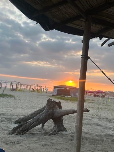 Captured at General Villamil Playas, Ecuador, the scene displays a sunset viewed from what appears to be a beachside structure with a thatched roof. A wooden support beam intersects the view, obscuring parts of the setting sun. A gnarled piece of driftwood rests on the sandy beach. In the background, beach umbrellas and temporary structures can be seen, with a few indistinct figures of people. The sky exhibits a gradient of colors typical of sunset, from yellows and oranges near the horizon to greys and blues higher in the sky. The foreground shows parts of the structure, with straw roofing elements and a supporting wooden post. A rope is tied to the post.