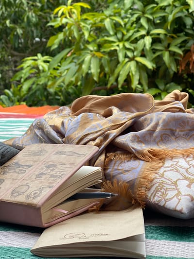The image shows an outdoor scene, potentially in Baan Kai, Thailand. Lush green foliage occupies the left side. On the right, a patterned silk cloth, predominantly in shades of gold and blue-gray, drapes over a textured surface with orange, green, and white stripes. Resting on the cloth are two open notebooks. One notebook is pink with anatomical drawings on the cover. A black pen is inserted in the pages of the notebook. The other notebook has a tan cover. The scene is composed to suggest a personal, reflective setting.