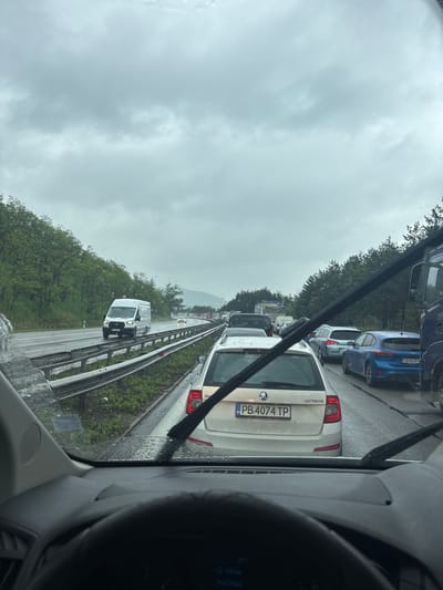 The image shows a traffic jam on a highway near Gorna Vasilitsa, Bulgaria. It is a cloudy day. Cars are lined up bumper to bumper in the right lane of the highway. A white Skoda Octavia with license plate PB 4074 TP is visible in the foreground. A white van is traveling in the left lane. The view is from inside a vehicle, looking through the windshield. Raindrops are visible on the windshield. Trees line the side of the highway.