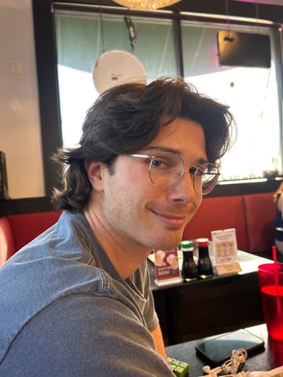 A male individual is seated at a table inside what appears to be a restaurant during daylight hours. He is positioned slightly off-center to the left, facing towards the viewer with a faint smile. The individual has dark, wavy, medium-length hair, wears silver-framed hexagonal eyeglasses, and exhibits light stubble with a mustache. He is dressed in a faded grey t-shirt. To his right, a blurry figure is partially visible seated in a red booth.
On the table in front of the male subject are multiple objects: a plate holding several small, green, rectangular food items; a dark-colored smartphone lying face down alongside white wired earbuds; two small condiment bottles, one with a green cap and one with a red cap; a small display stand with two white cards, one of which includes the text "SOJU"; and a translucent red plastic cup with a red straw containing a clear liquid.
The background features red booth seating and large windows that illuminate the scene with bright daylight. A white, circular object with a small protrusion is visible through the windows. The restaurant interior includes dark wall panels, and a portion of a decorative light fixture is visible overhead. An electrical wall outlet is present on the far-left wall. The specific location of this restaurant within Lomita, United States of America, is not discernible from the image.