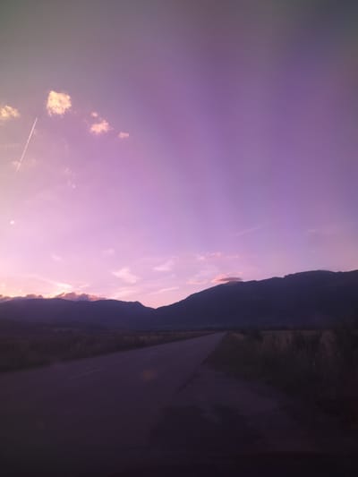 The image depicts a road stretching into the distance, flanked by fields of dry grass and bordered by a dark silhouette of mountains under a vivid purple and pink sky. A contrail from an aircraft is visible in the upper left corner of the sky, along with scattered clouds. The lighting suggests either dawn or dusk. The location is identified as Manolovo, Bulgaria. There are no people or animals visible in the frame. The road appears to be asphalt with a broken white line marking the center. The overall mood is serene and expansive.