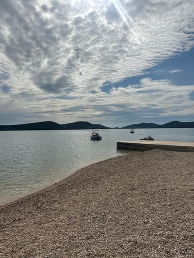 The image depicts a coastal scene in the city of Betina, Croatia. In the foreground, a light-colored pebble beach slopes gently into a body of water, occupying the lower third of the frame. To the right, a concrete pier or dock extends into the water.
The midground features calm water with significant sunlight glinting and shimmering across its surface, creating a bright path. Three vessels are visible: a white motorboat with a cabin is anchored in the left-center, a smaller grey motorboat is further back towards the right, and a black jet ski is moored alongside the concrete pier on the right. Several dark, circular mooring buoys are sparsely distributed across the water.
In the background, multiple tree-covered islands or landmasses with varied elevations stretch across the horizon. The sky occupies the upper two-thirds of the image, characterized by a mix of altocumulus clouds, with some patches of clear blue sky, particularly towards the upper right. Bright sunlight emanates from the upper-left corner, with visible rays piercing through the clouds. No people are visible, and no active actions are depicted.