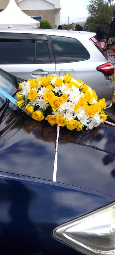 This image depicts an outdoor celebratory scene in Sigona ward, Kenya, dominated by a dark blue car adorned with a vibrant floral arrangement on its hood. The arrangement is a prominent display of numerous yellow roses interspersed with clusters of small white chrysanthemums, secured with a light blue ribbon on the left and a white ribbon patterned with small red designs further on the hood. The car's highly reflective surface shows a clear blue sky with white clouds.
Behind the decorated car, a silver vehicle is partially visible, showing its rear section and tinted side windows. Several individuals are present in the background near the silver car, though their faces are not clearly discernible. One person on the far right appears to be dressed in traditional or formal attire featuring gold embroidery.
The setting includes a white event tent on the left side and a light-colored building in the background, suggesting a venue for a gathering or ceremony. Green foliage and bushes contribute to the outdoor ambiance. The bright daylight indicates a sunny or partly cloudy day, enhancing the festive mood. The presence of the decorated car, often used as a bridal vehicle, strongly suggests a wedding or a similar significant celebratory event. No visible text is present in the image.