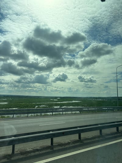 The image captures a view from a moving vehicle on a highway, likely in the vicinity of Menzelinsk, Russia, given the landscape. A two-lane highway with a metal guardrail dominates the foreground. Beyond the highway, a vast expanse of green marshland dotted with bodies of water stretches into the distance. The sky above is filled with dramatic, gray and white clouds, suggesting a dynamic weather pattern. A single lamppost is visible on the right side of the image. The overall scene conveys a sense of travel through a rural or semi-rural landscape.