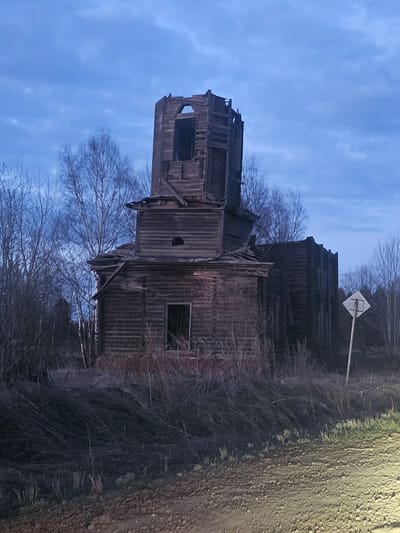 The image captures a large, derelict wooden structure, possibly an old church or a substantial traditional building, set against a soft, twilight blue sky. The building is constructed from dark, weathered wood planks and exhibits significant signs of decay, with missing sections, exposed internal beams, and crumbling elements, particularly in its multi-tiered and tower-like upper portions. Several window openings are visible, appearing empty.
Bare, leafless trees and dry brush encircle the structure, contributing to an atmosphere of abandonment and history. On the right side of the frame, a dirt or gravel road runs vertically, indicating a rural or remote setting. Behind the main building, a white diamond-shaped sign mounted on a pole is partially visible through the branches.
The sky is a gentle, diffused blue with light cloud formations, suggesting either early morning or dusk. The soft lighting casts no harsh shadows, accentuating the quiet and melancholic mood of the scene. No individuals are present, and no activity is occurring. The intricate wooden construction style is characteristic of traditional Russian architecture, aligning with the location context of Kizhi, Russia, renowned for its historic wooden buildings. There is no visible text in the image.