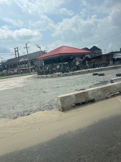 The image presents an outdoor street scene in what appears to be a semi-urban or developing area, consistent with the location context of Asubi, Nigeria. The weather is fair, with a bright sky featuring scattered white cumulus clouds, indicating daytime.
In the foreground, a paved road surface occupies the right side of the frame, partially covered by a layer of light-colored sand and dirt. A sturdy concrete curb or barrier, showing signs of wear and tear, runs parallel to the road on the left. Beyond the curb, there's a rough strip of ground mixed with gravel and dirt.
The mid-ground is dominated by a distinctive building featuring a prominent red sloped roof and walls adorned with a green, brown, black, and white camouflage pattern. Several dark, possibly cylindrical objects (resembling tires or drums) are positioned near this building. Further into the background, other buildings of varying sizes and designs can be seen, suggesting a mix of residential and perhaps commercial structures.
Numerous utility poles with a dense network of overhead electrical wires are visible on the left side, extending into the sky. No people are discernibly present in the scene, and there is no legible text visible on any signs or structures. The image captures a static moment of this street environment, with no particular activity or event taking place.