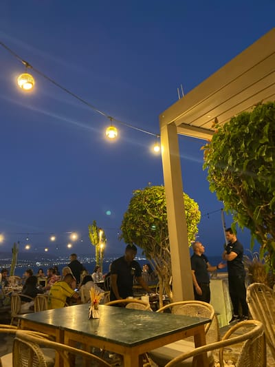 An outdoor dining area at dusk in Taghazout, Morocco, features tables with green tops and wicker chairs, illuminated by strings of spherical yellow lights. Two men in dark shirts are conversing near a beige pillar and a tree with lush green foliage. In the background, a distant city lights up against the dark blue sky, suggesting a coastal location. Several other patrons are seated at tables, engaged in dining and conversation, with more decorative lights strung overhead. The overall atmosphere is one of a lively evening gathering.