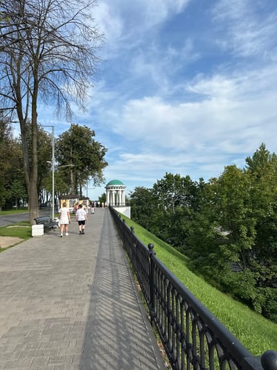 The image depicts a scenic park pathway in Grigor'evskoe, Russia, on a clear day. The pathway is paved with gray rectangular stones and bordered by a black wrought-iron fence on the right, which separates it from a steep, grassy incline covered in lush green grass. Beyond the fence and incline, dense green trees form a natural backdrop. On the left side of the pathway, a paved road is visible, lined with trees. A dark-colored park bench with white stone bases sits near the beginning of the pathway. Further along the path, a white gazebo with a green dome is visible, standing prominently against the blue sky with scattered white clouds. Several people are walking on the pathway; in the foreground, a woman in a white dress and a person in a white t-shirt and black shorts are walking away from the camera. Other individuals are dispersed further down the path, some near small kiosk-like structures. The overall scene suggests a public recreational area enjoying pleasant weather.
