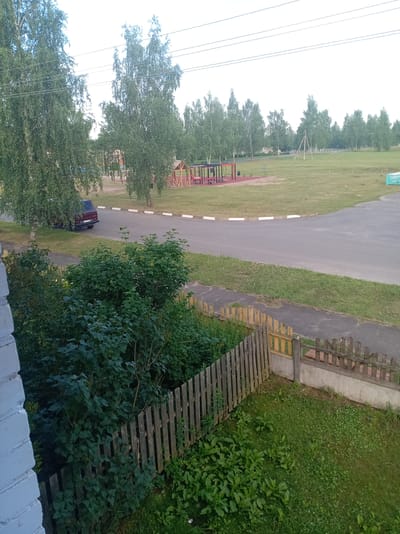 The image depicts an outdoor scene on a clear day, likely in a park or recreational area within a rural or suburban setting, possibly in the city of Пралетарскі сельскі Савет, Belarus. In the foreground, a weathered wooden fence runs diagonally across the frame, bordered by lush green grass and dense shrubbery. Beyond the fence, a paved pathway leads to a road.
Further into the scene, a red vehicle is parked near the roadside, partially obscured by a large birch tree. Across the road, a children's playground with a red and wooden structure is visible. The playground is situated on a sandy area surrounded by grass. In the background, a line of trees forms a natural barrier, with more trees and open grassy fields extending towards the horizon. Overhead, power lines are strung across the sky. The overall impression is of a quiet, well-maintained public space.