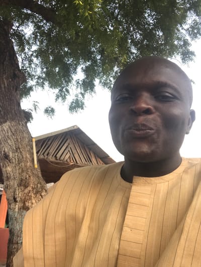 This image captures a bald, dark-skinned male subject in a close-up, front-facing perspective, situated in Ugwolawo, Nigeria. The subject wears a light tan garment featuring vertical pinstripes and a distinctive collarless neckline with a pleated placket. His lips are pursed as he looks directly at the camera. To the left, a portion of a tree trunk with lighter markings is visible, extending upwards into green foliage that partially covers a bright, diffused sky. Behind the subject's left shoulder, a rustic wooden structure composed of slatted timber elements is partially observable. The scene is outdoors and appears to be illuminated by natural daylight.